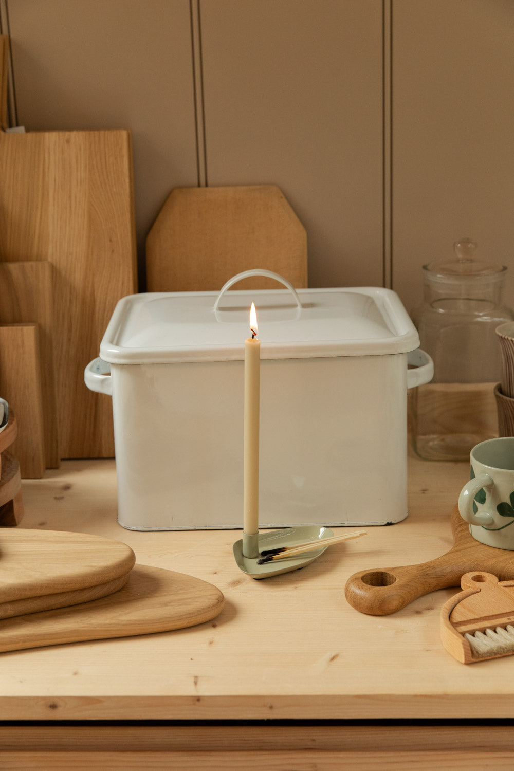 Styled pine kitchen counter with white enamel bread bin, stacked oak chopping boards, lit cream taper candle, white enamel kettle and assorted wooden homeware against taupe panelled wall