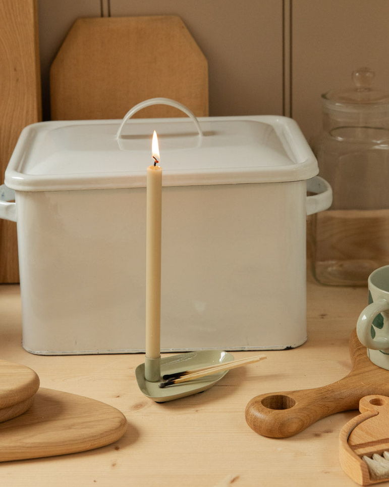 Styled pine kitchen counter with white enamel bread bin, stacked oak chopping boards, lit cream taper candle and assorted wooden homeware against taupe panelled wall