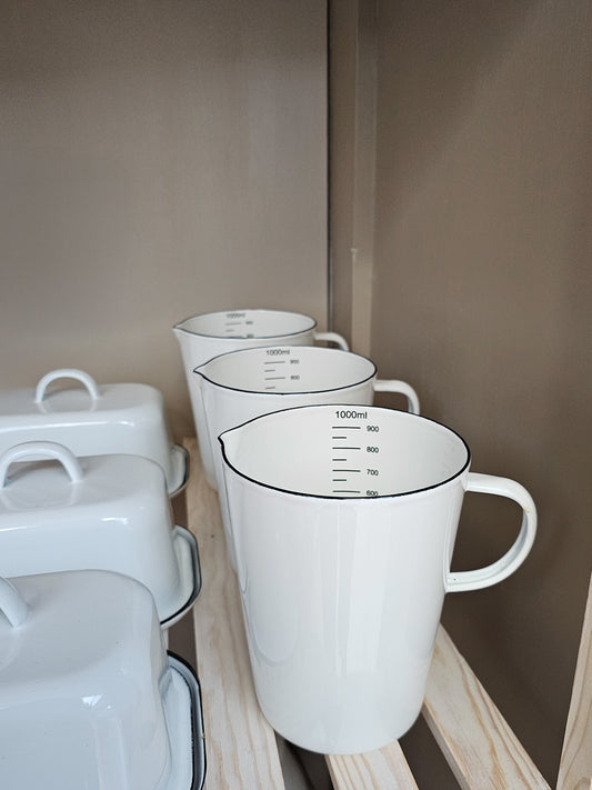 A white enamel measuring jug with black markings and a handle, displayed in a kitchen setting.
