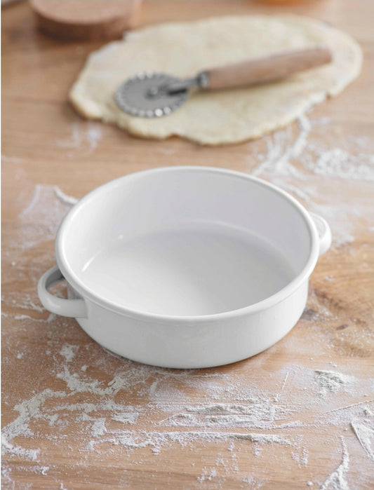 A white enamel pie dish with two handles, placed on a wooden surface with flour on it, next to uncooked dough and a rolling pin.