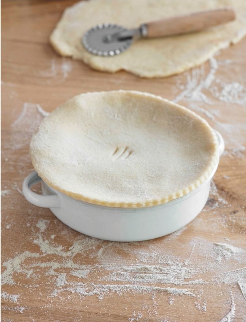 A white enamel pie dish with two handles, placed on a wooden surface with flour on it, next to uncooked dough and a rolling pin.