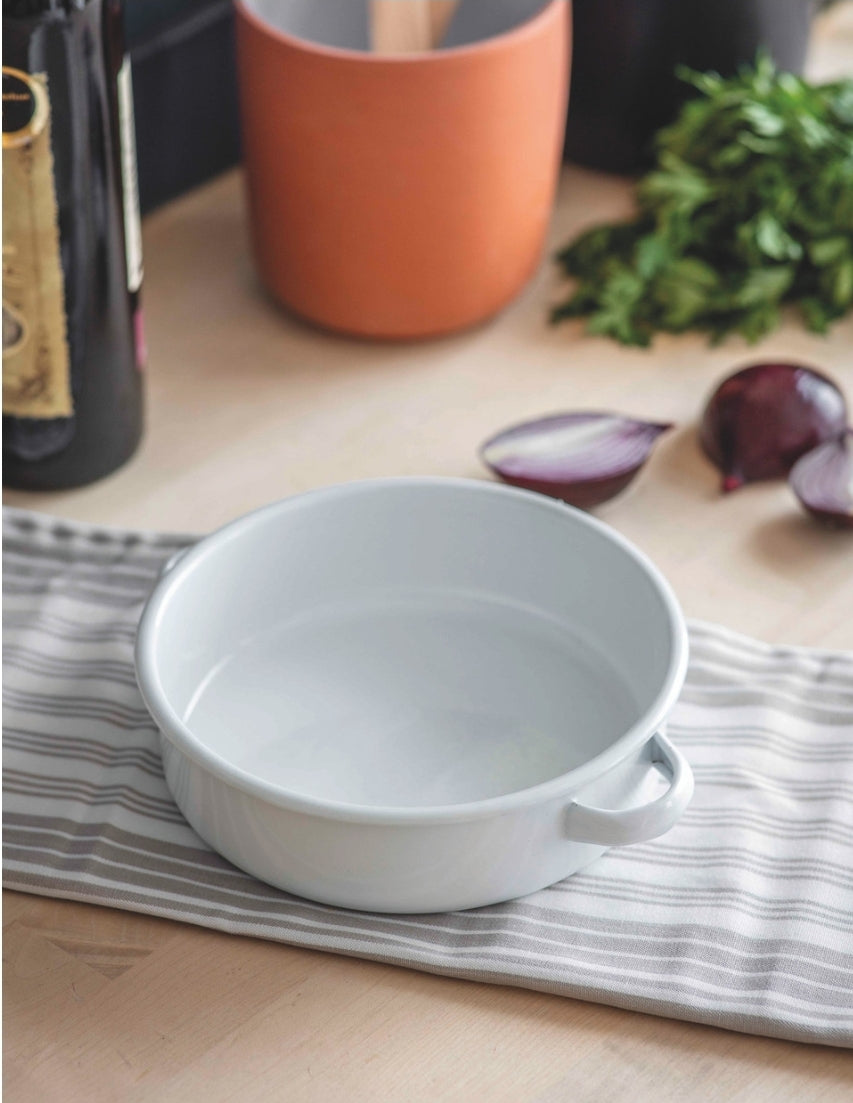 An empty white enamel pie dish with two handles, placed on a tea towel on a wooden surface.