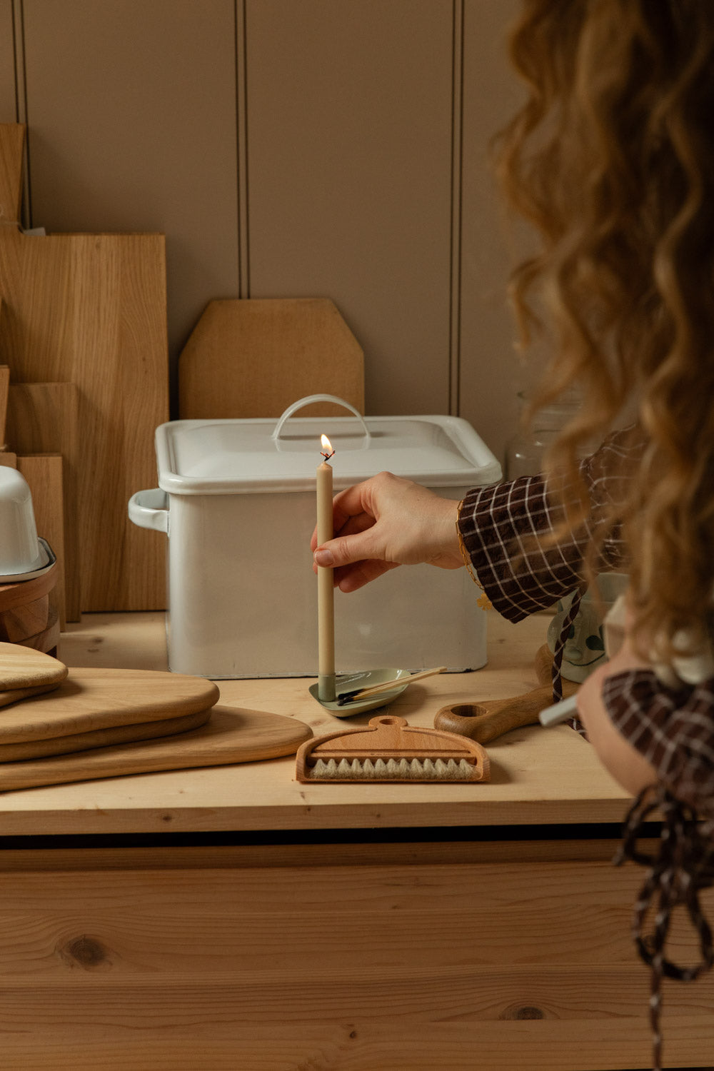 Woman lighting a cream taper candle in a sand coloured metal holder on a pine kitchen counter, surrounded by oak chopping boards, a white enamel storage box and wooden homeware