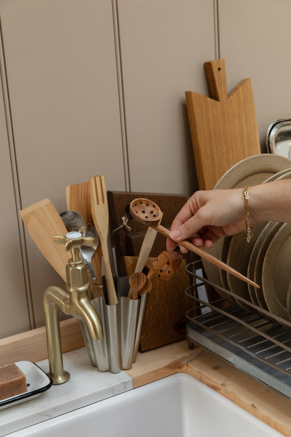 Hand taking a wooden spoon from a steel pot of wooden kitchen utensils beside a brass tap
