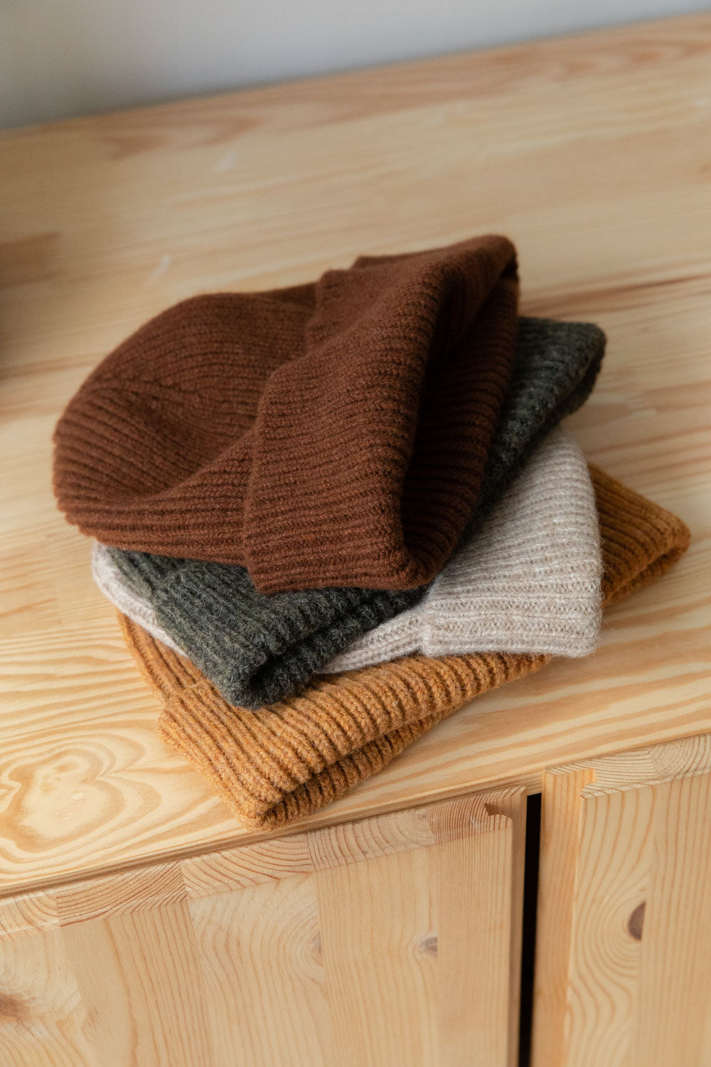 Stack of four wool beanies in brown, green, oatmeal and orange on a pine cabinet