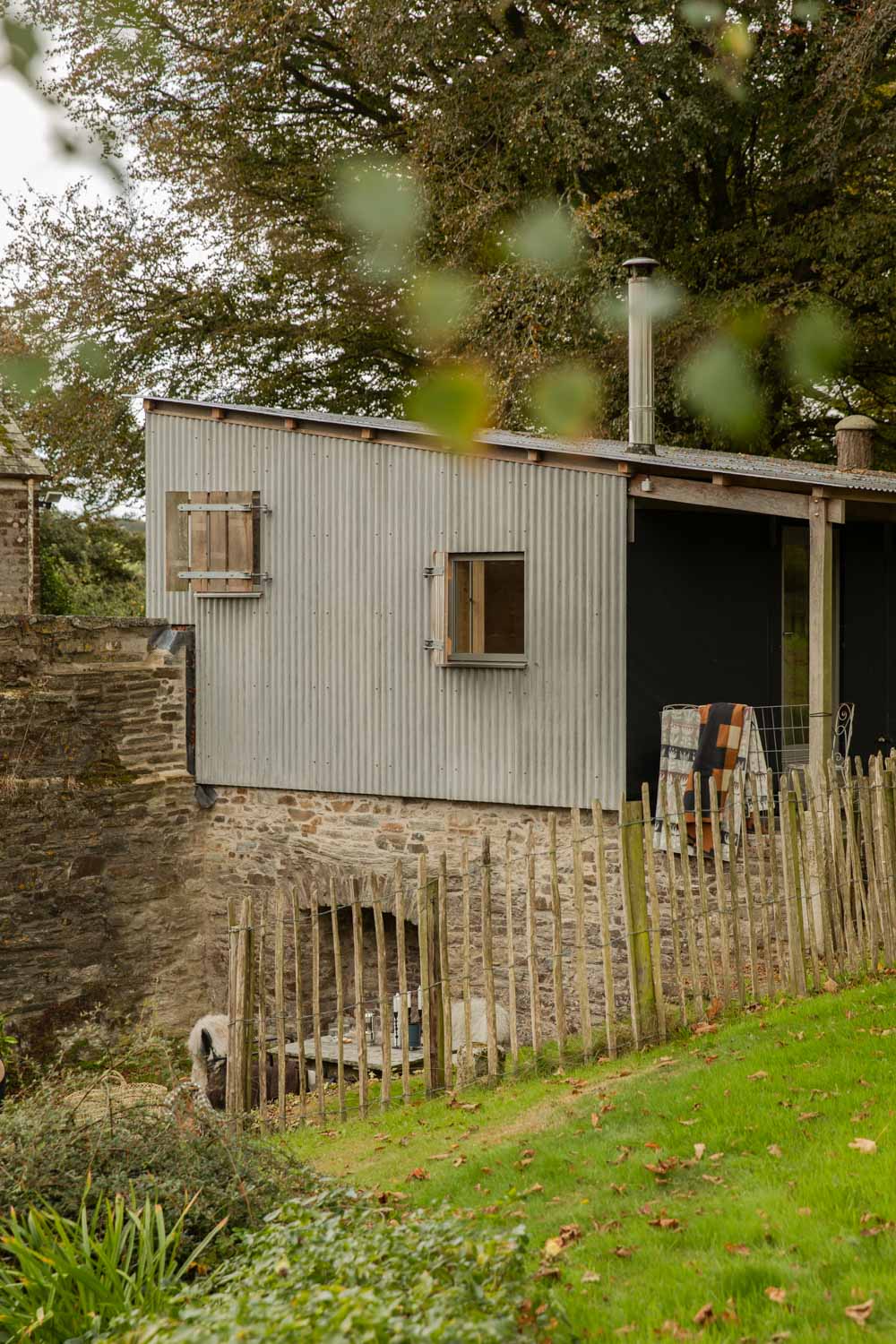 Tin-roofed cabin in an autumn garden with wool blankets airing on the veranda rail behind a chestnut paling fence