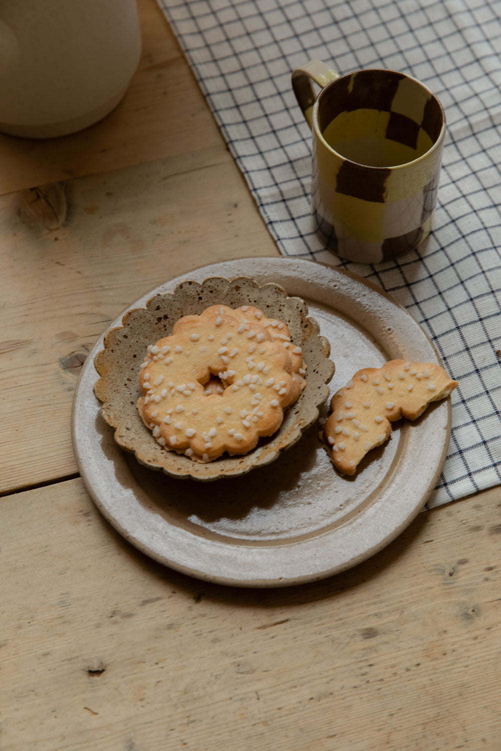 Yellow and brown check mug beside biscuits on a scalloped dish and speckled stoneware plate, on a grid linen placemat