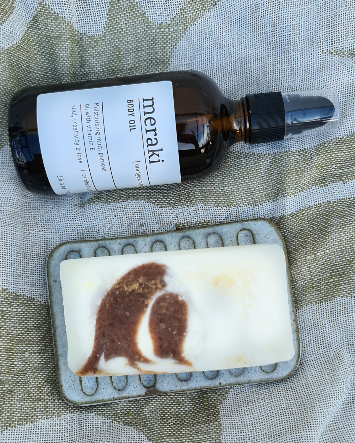 Illustrated bar of soap on a rustic ceramic soap dish beside an amber dropper bottle on linen