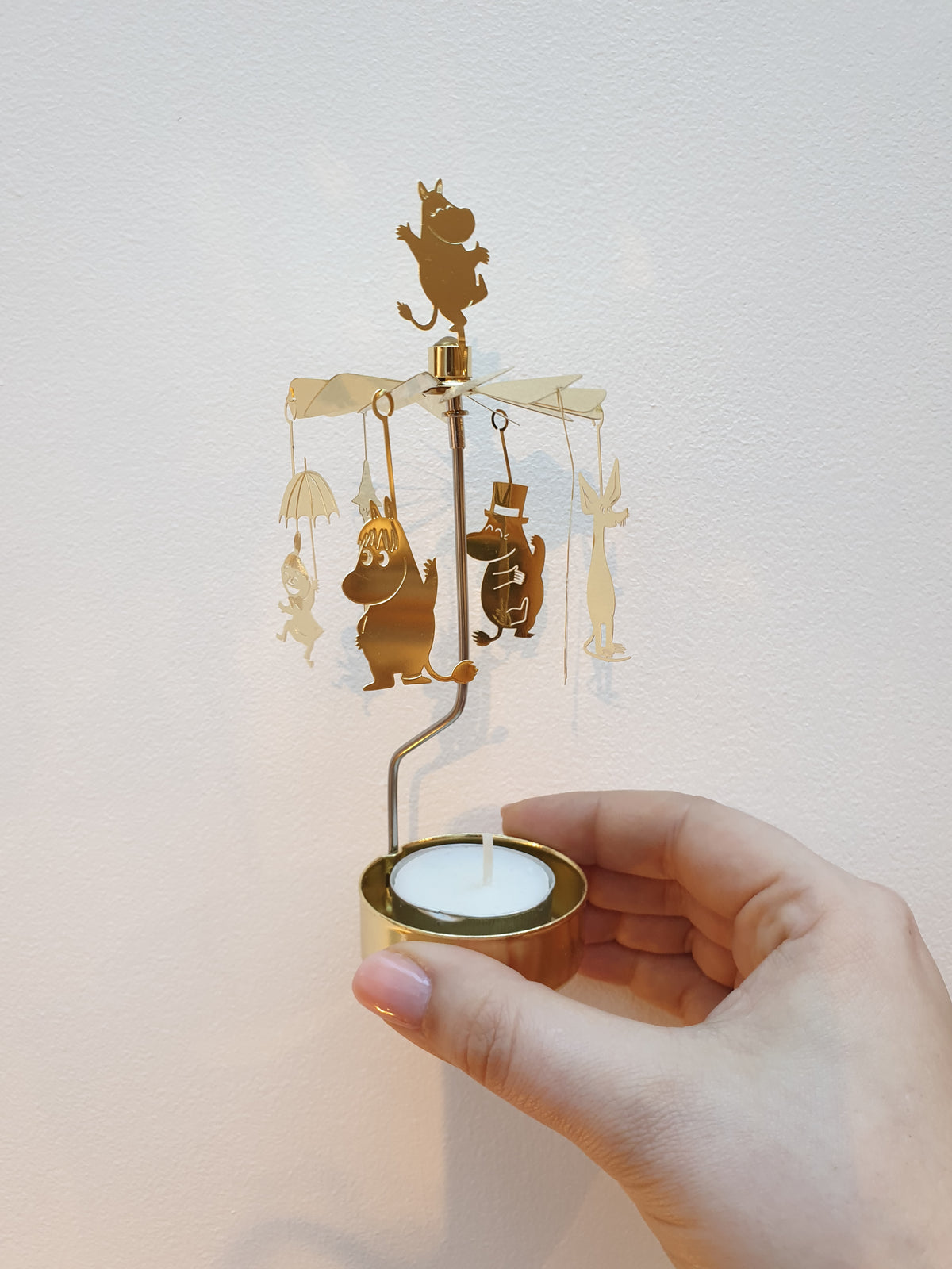 Hand holding a gold Moomin Änglaspel, showing the Moomin figures hanging from its arms