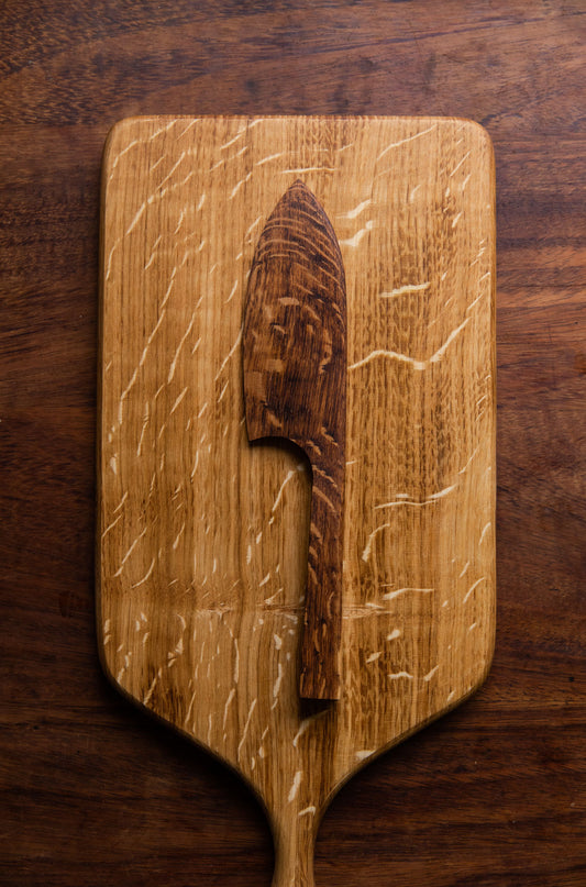 A brown English Oak cake knife rests on top of a serving board, placed on a dark wooden worktop.