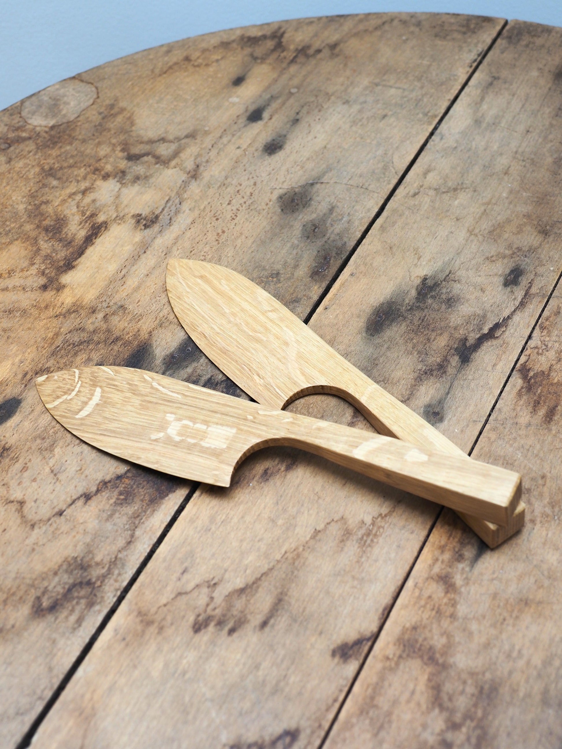 Two English Oak cake knifes resting on top of each other on a wooden table.