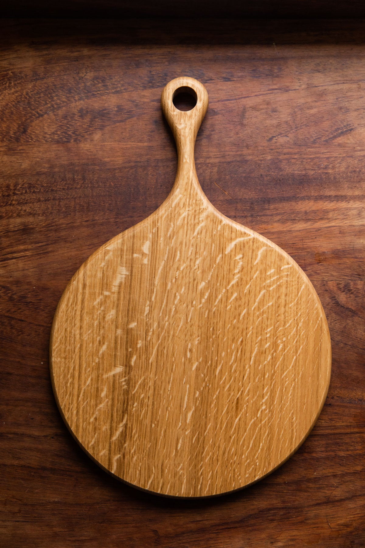 An English Oak pizza board is resting on a dark brown wooden tabletop. Taken from a birds eye view.