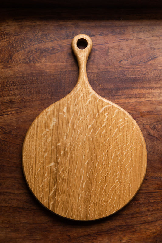 An English Oak pizza board is resting on a dark brown wooden tabletop. Taken from a birds eye view.