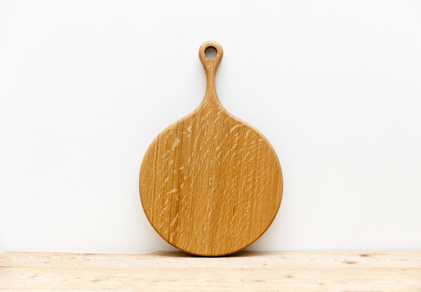 An English Oak pizza board is shown resting against a white wall, sitting on a light wooden table.