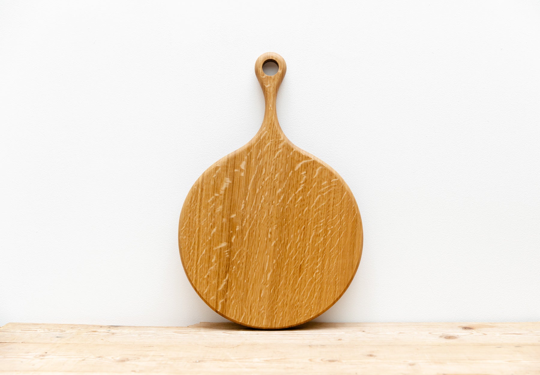 An English Oak pizza board is shown resting against a white wall, sitting on a light wooden table.