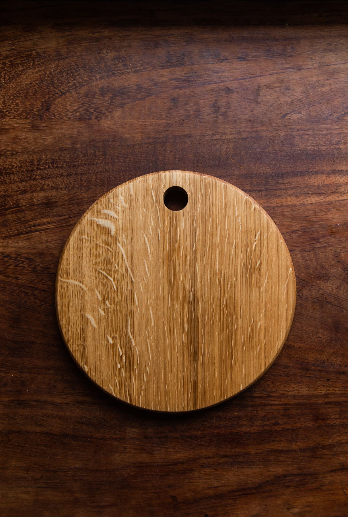 A round, English oak wood board with a hole at the top, placed on a dark wooden surface.