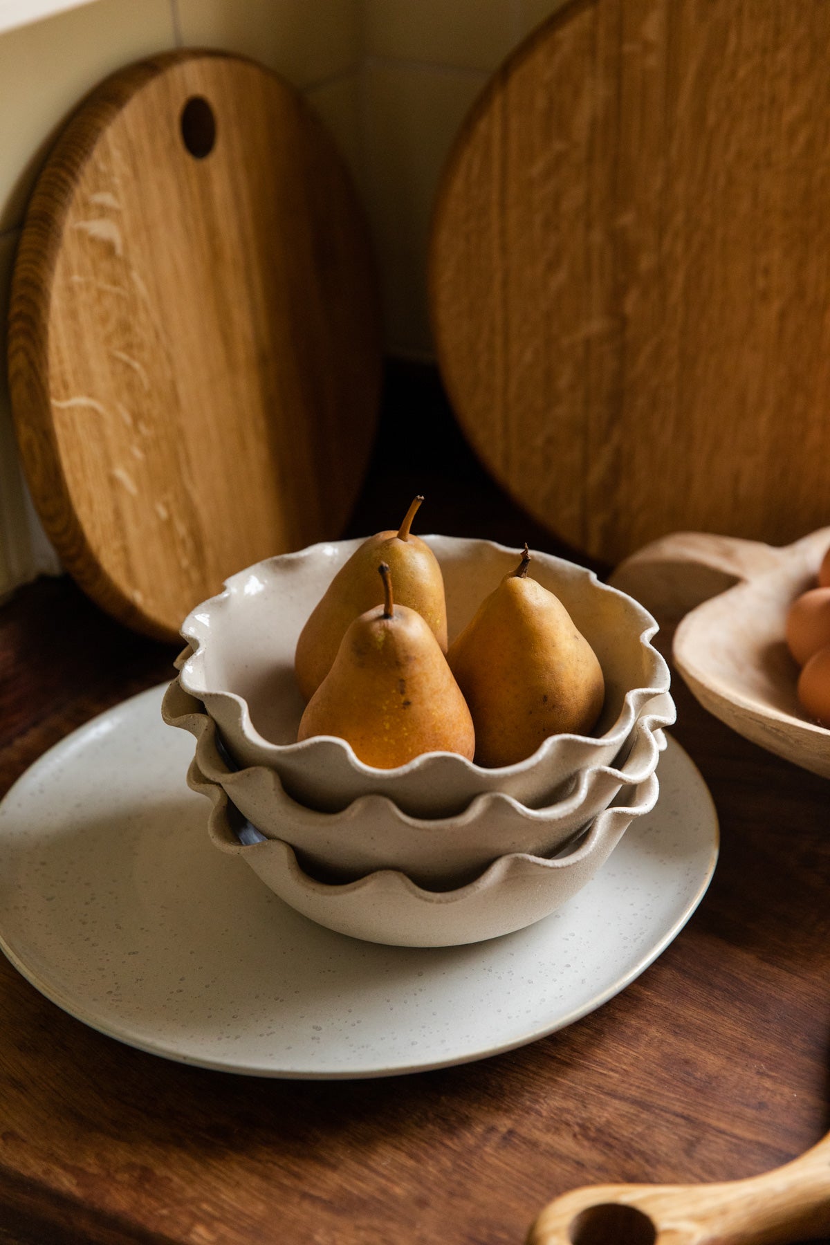 A round, English oak board with a hole at the top is seen leaning against a wall in the background of a kitchen. The focal point of the image is a pile of bowls holding three pairs.