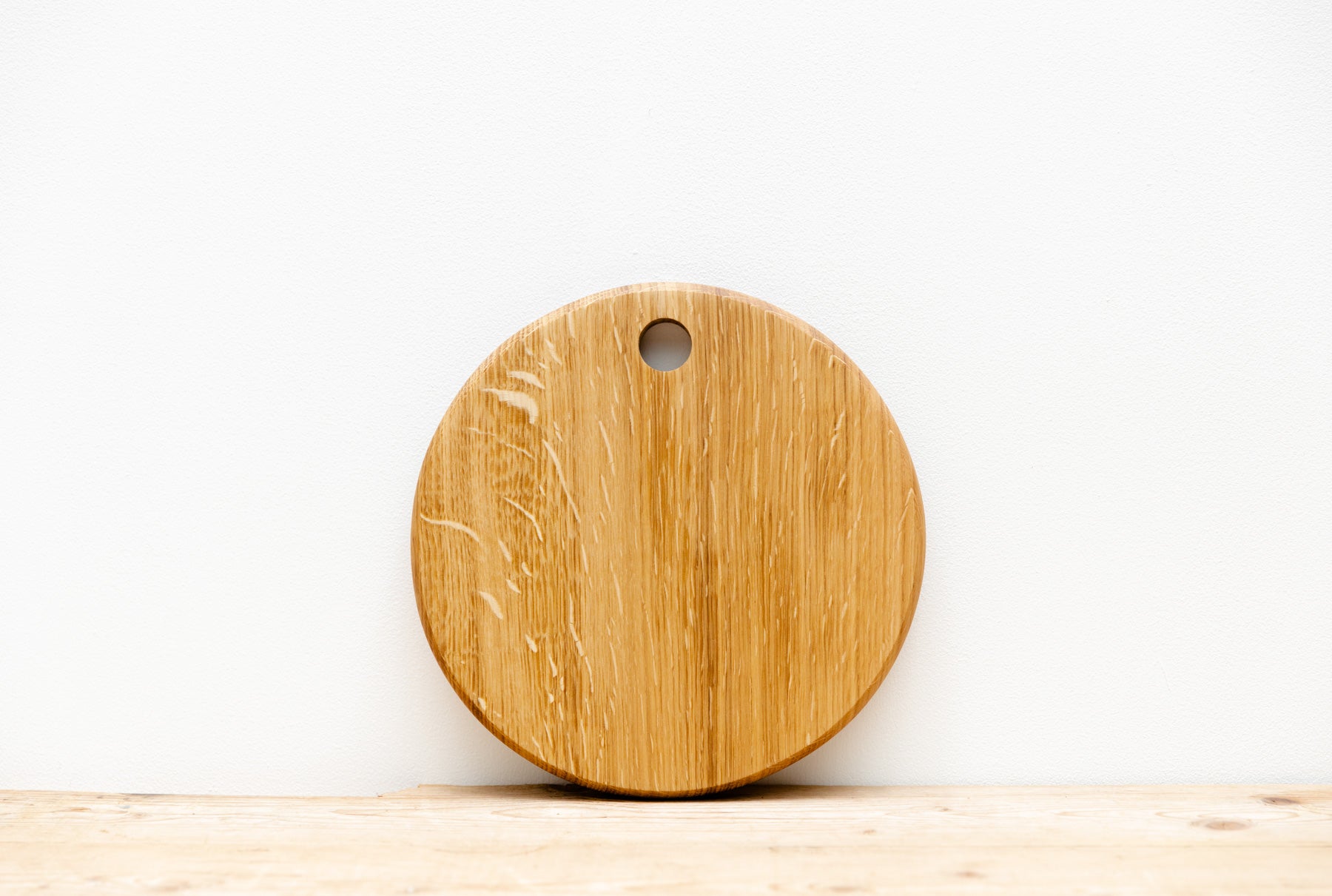 A round, English oak wood board with a hole at the top, placed on a wooden surface, leaning against a white wall.