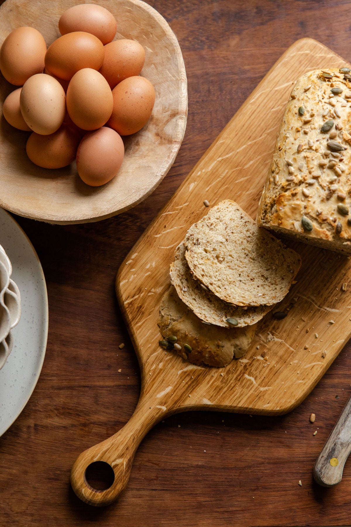 A serving board made from English Oak resting on a wooden table. There is a cut up loaf of seeded bread placed on the board and a bowl of eggs placed nearby.