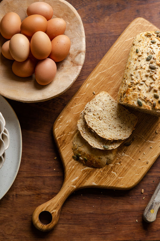 A serving board made from English Oak resting on a wooden table. There is a cut up loaf of seeded bread placed on the board and a bowl of eggs placed nearby.