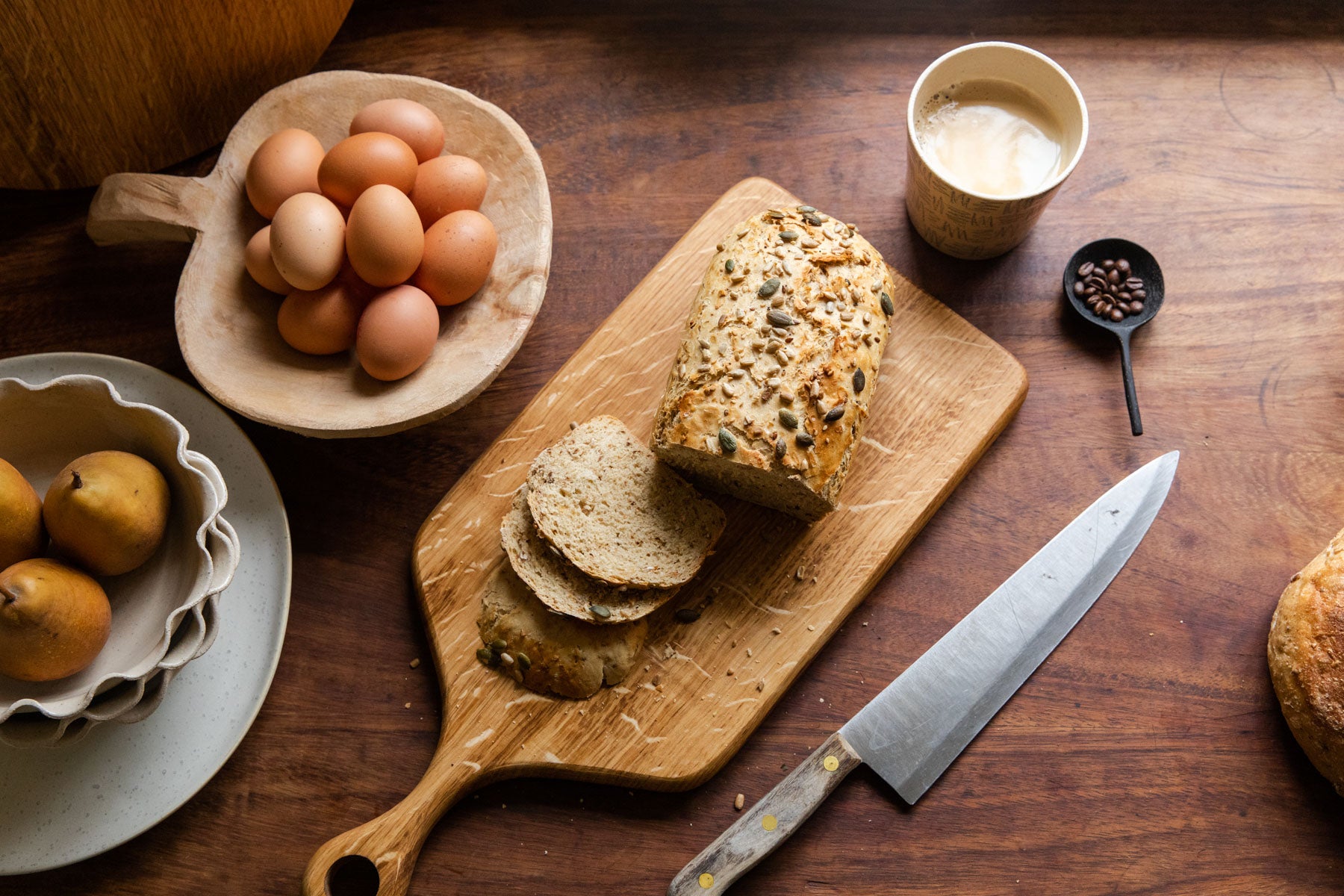 A table display showing the English Oak serving board with a loaf of cut up seeded bread on top of it. Close by on the table there is a large knife, a cup of coffee, a coffee scoop with coffee beans inside of it, a wooden bowl filled with eggs and another stack of bows with pears inside. 