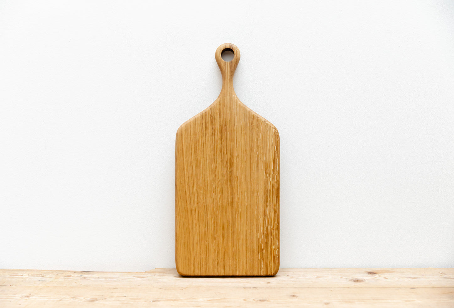 An English Oak serving board leaning up against a white wall, sitting on  wooden worktop.