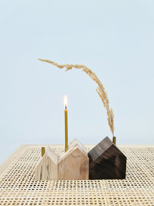 Trio of wooden house shaped candle holders with brass tube chimneys, one with small candle, one with stem of dried grass in.