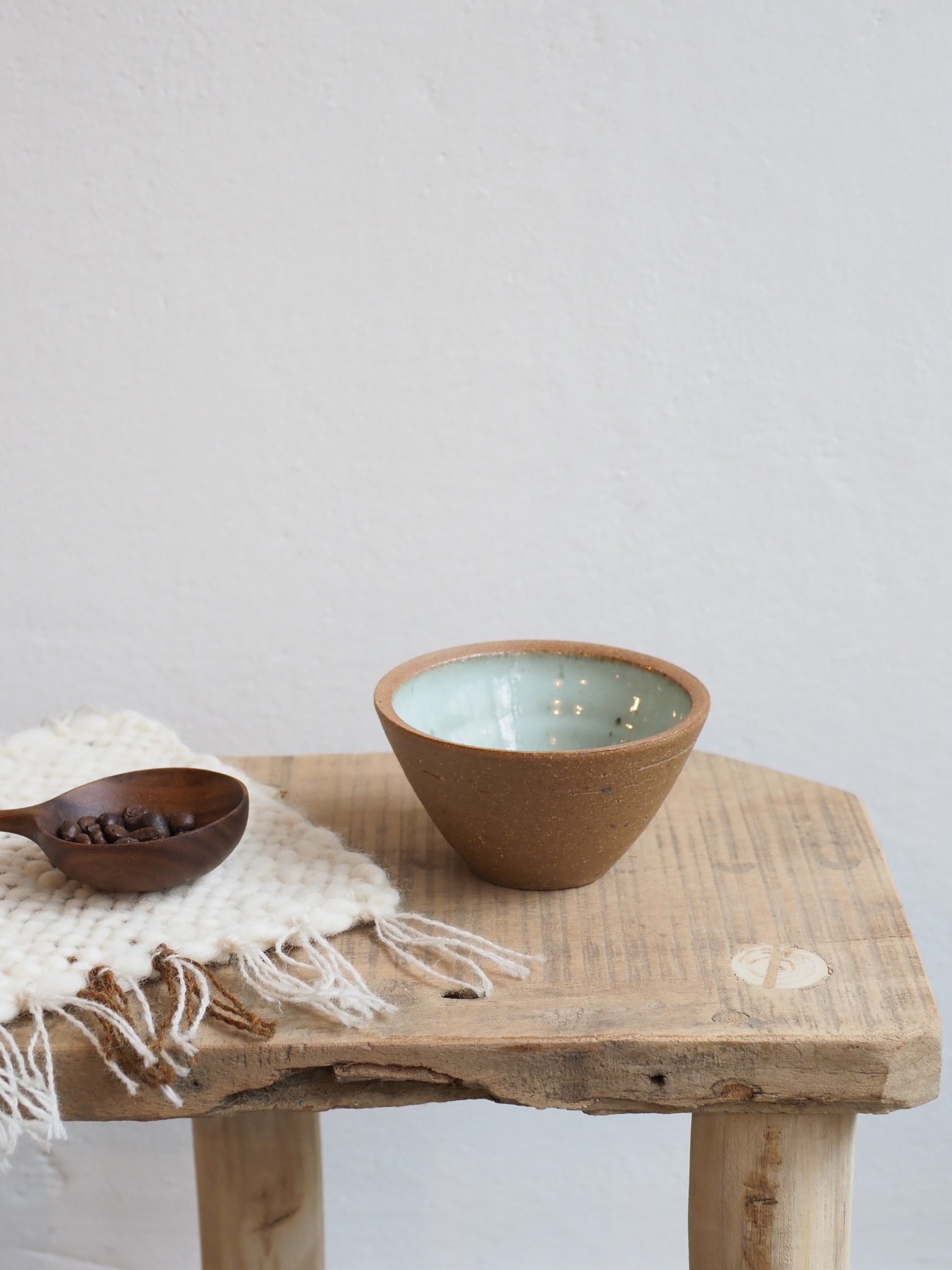 Leach Pottery small prepping bowl with Dolomite glaze inside on a wooden table. Also on the table is a wooden coffee scoop with coffee beans inside and a handmade woven coaster.