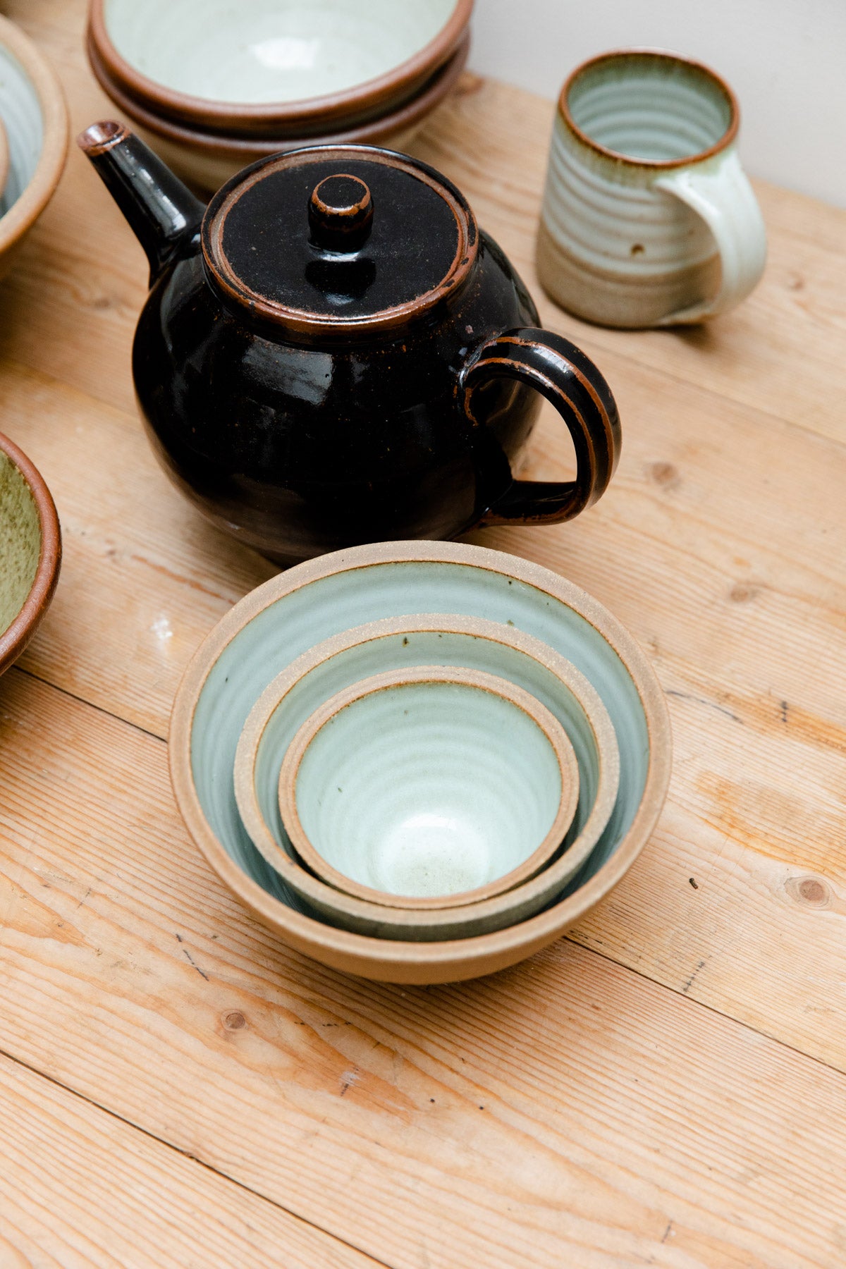 Three Leach Pottery prepping bowls with Dolomite glaze inside on a wooden table. The bowls are nested inside one another, next to a black Leach Pottery teapot, and mug.
