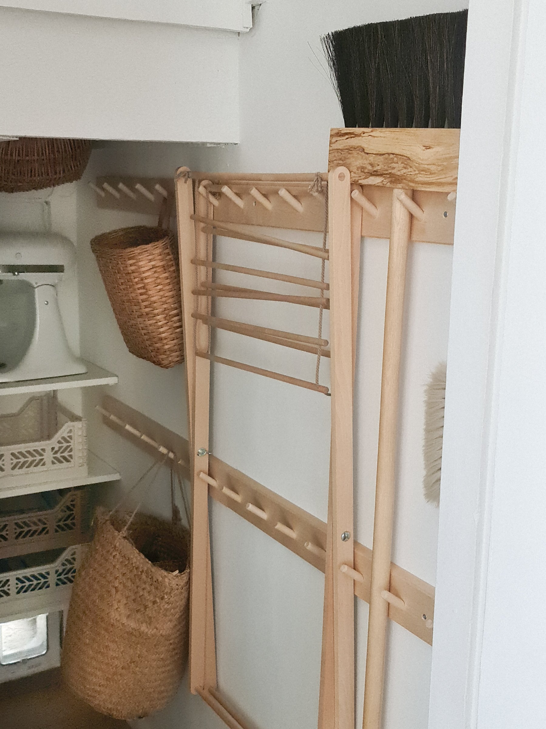 A peg rail rack made of birch wood with multiple hooks, mounted on a wall in an under stairs cupbaord. It's holding various items such as a broom, wooden clothes airer and basket.