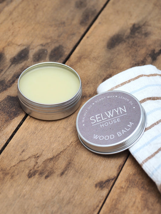 A small open tin of wood balm next to its screw-top lid, with a white cloth on a wooden surface. The tin has a label that reads 'Selwyn House Wood Balm'.