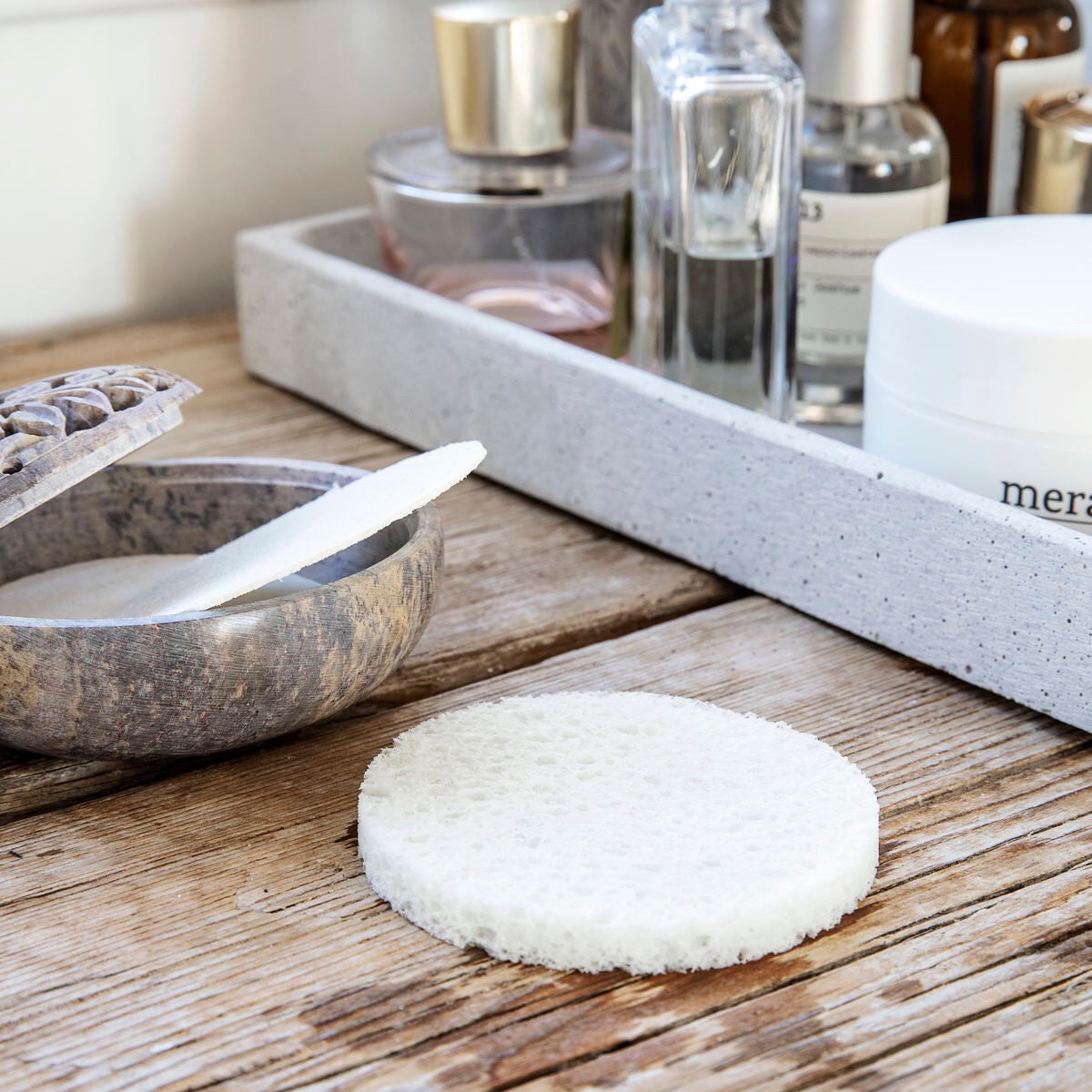 An expanded facial cleaning sponge sits on a wooden surface in a bathroom setting. 