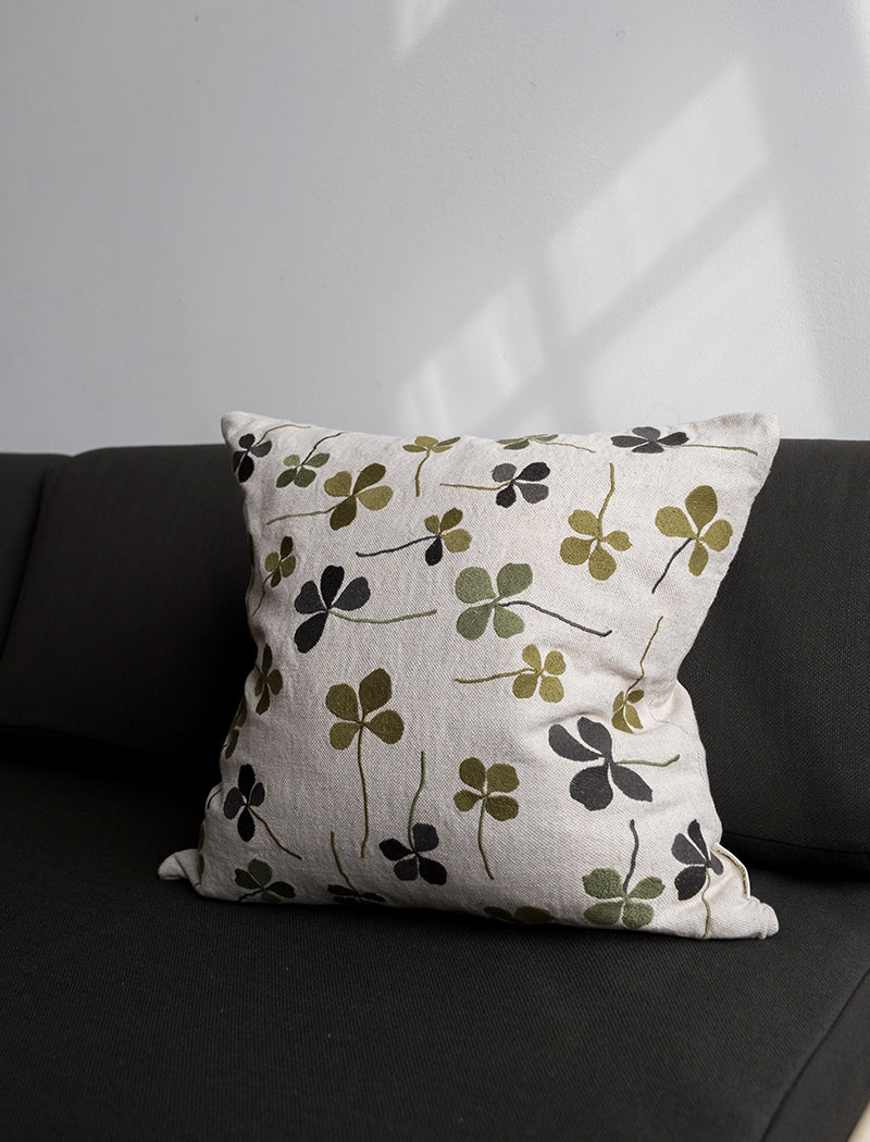A linen coloured cushion with a green clover embroidery pattern on a black sofa.