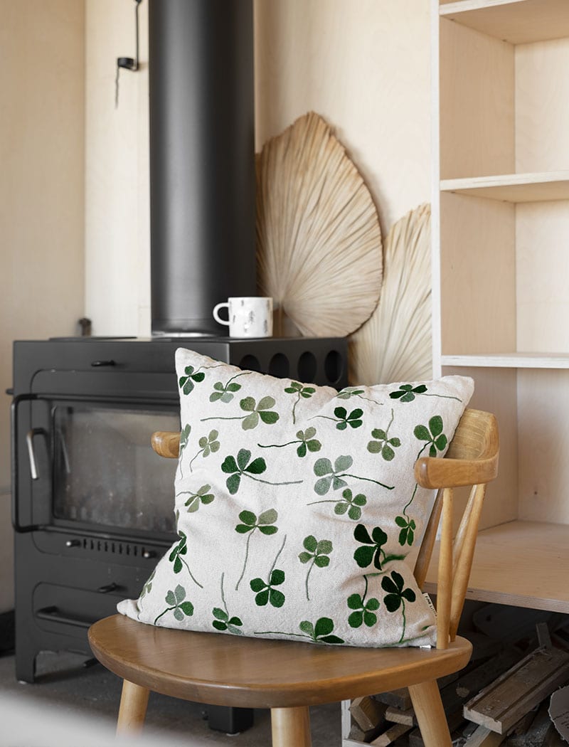 A linen coloured cushion with a green clover embroidery pattern on a wooden chair next to an indoor fireplace.