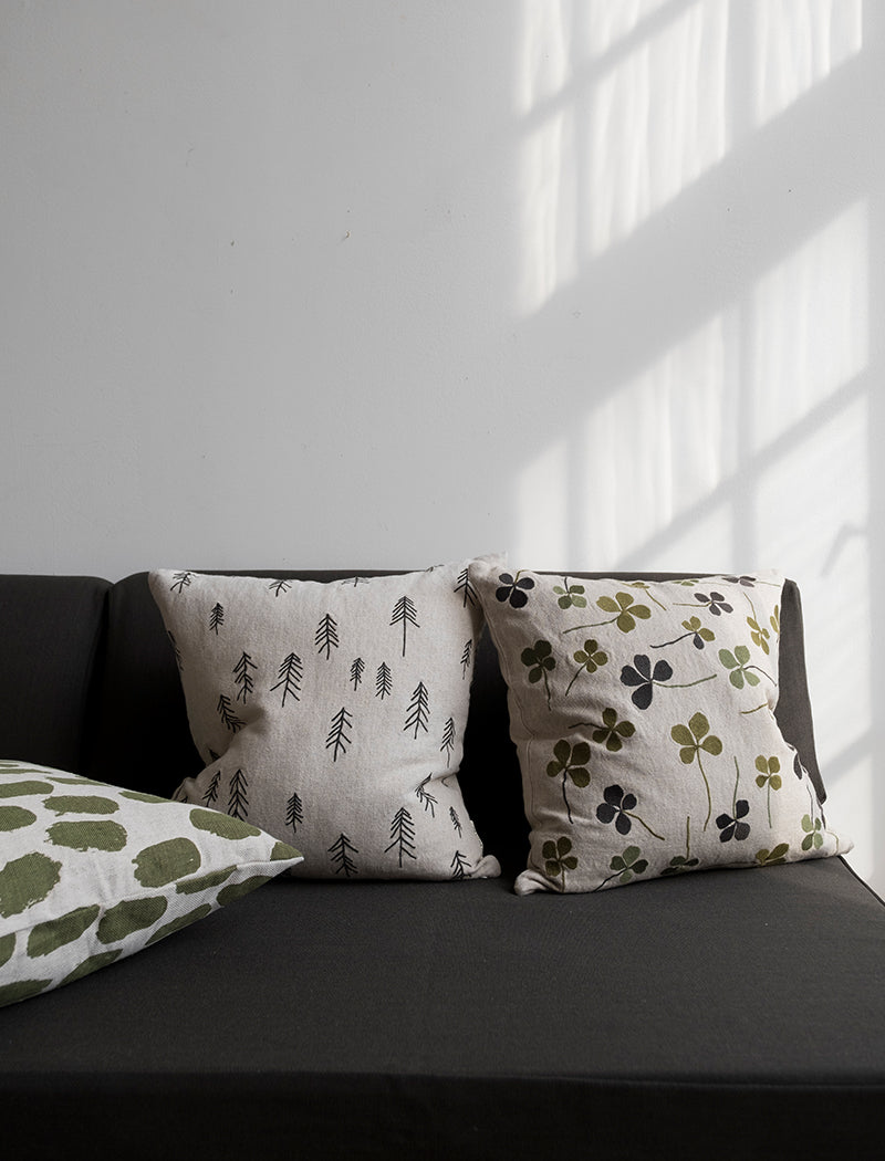 A linen coloured cushion with a green clover embroidery pattern on a black sofa next to other cushions.