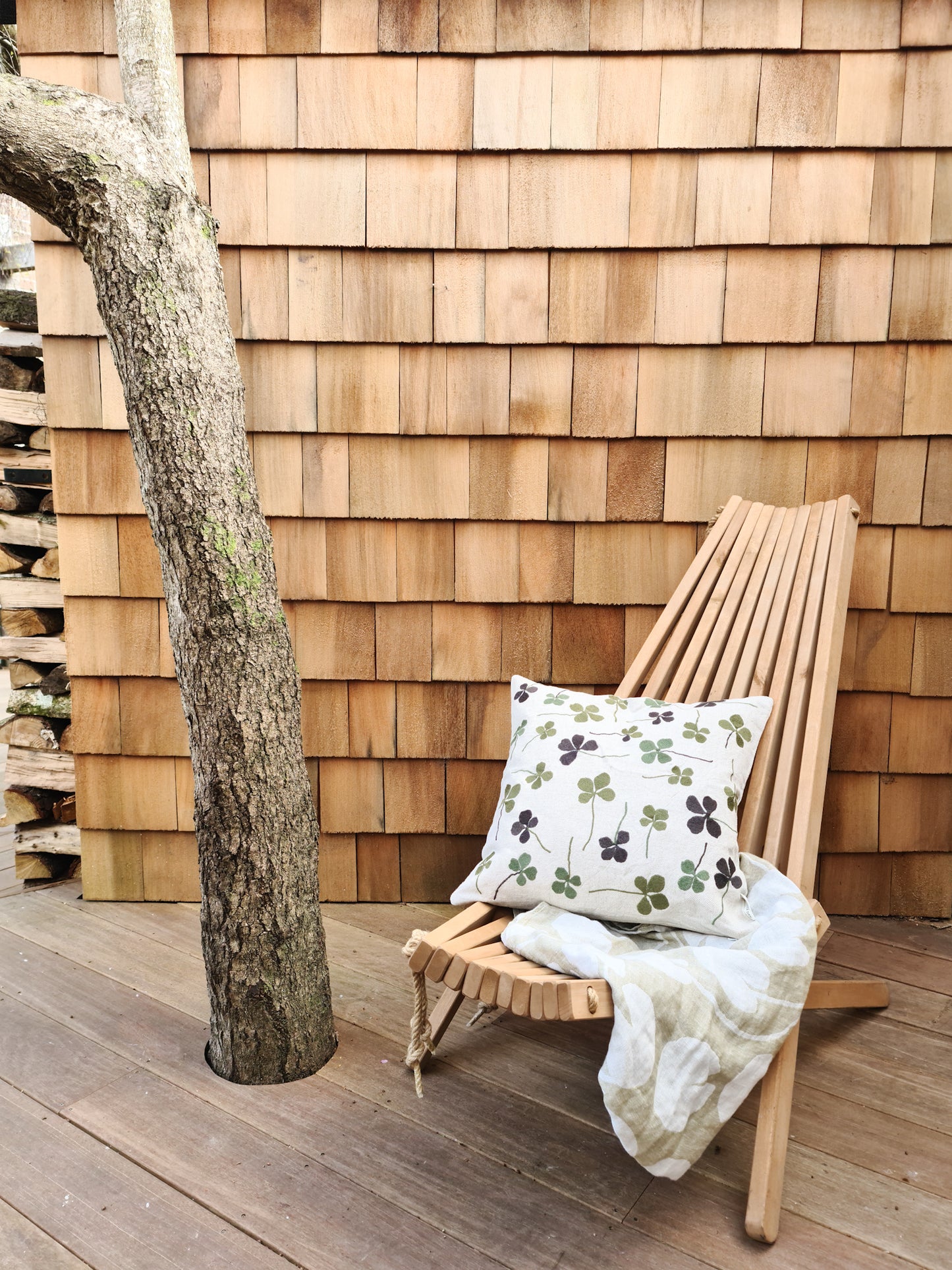 A linen coloured cushion with a green clover embroidery pattern on a wooden chair in front of a garden shed and tree.