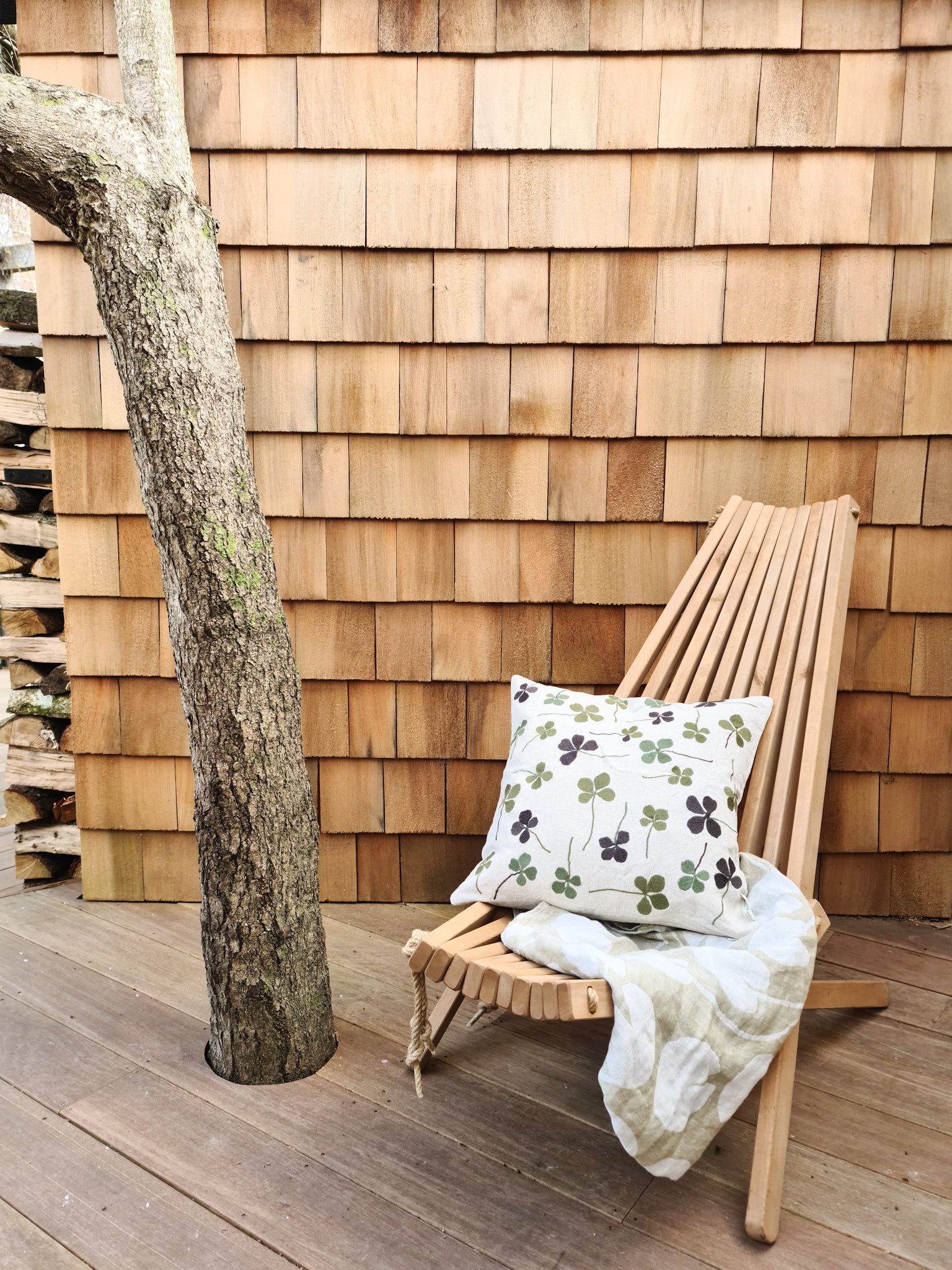 A linen coloured cushion with a green clover embroidery pattern on a wooden chair in front of a garden shed and tree.