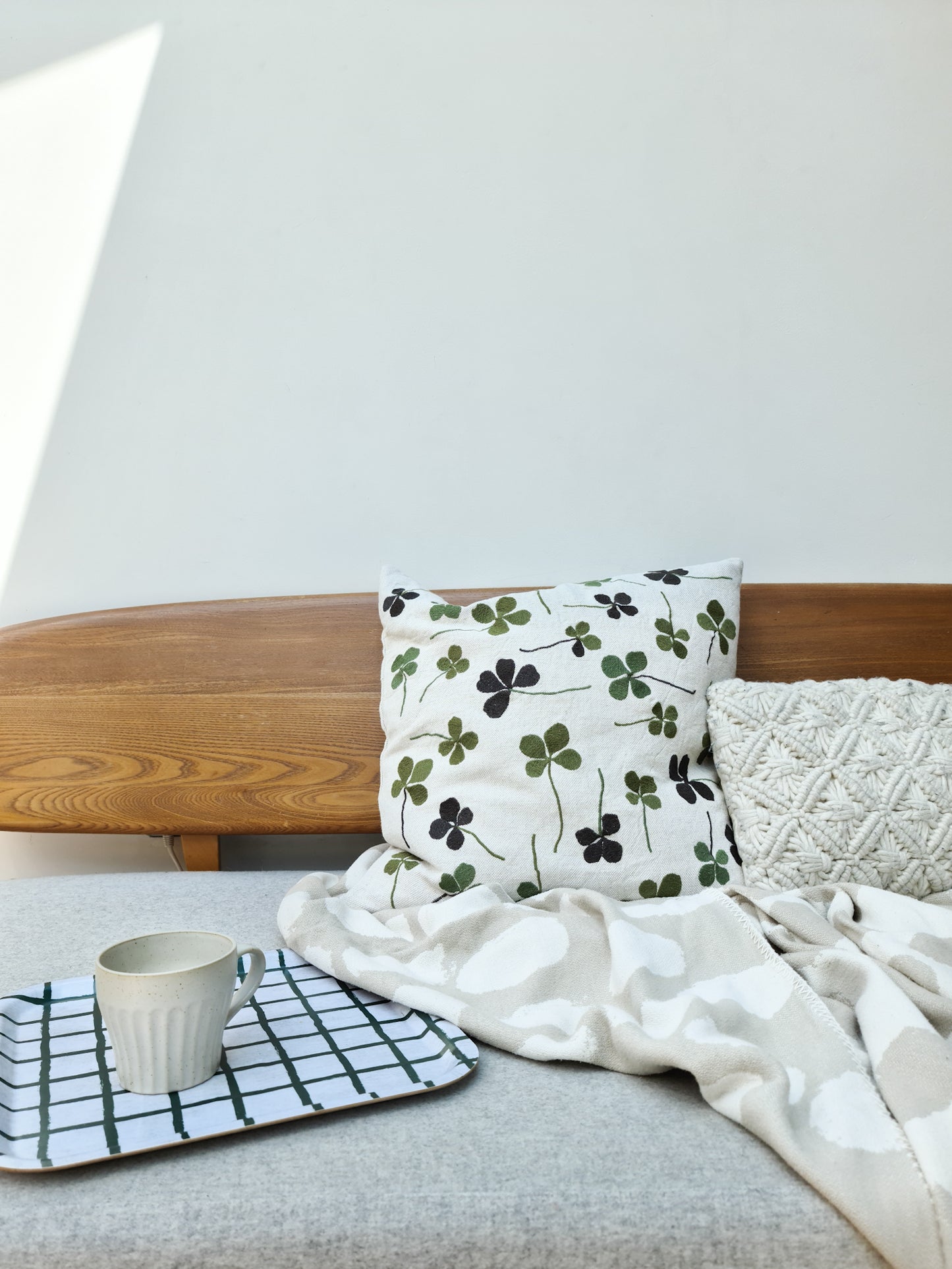 A linen coloured cushion with a green clover embroidery pattern on a sofa, next to a blanket and tray with a mug on it.