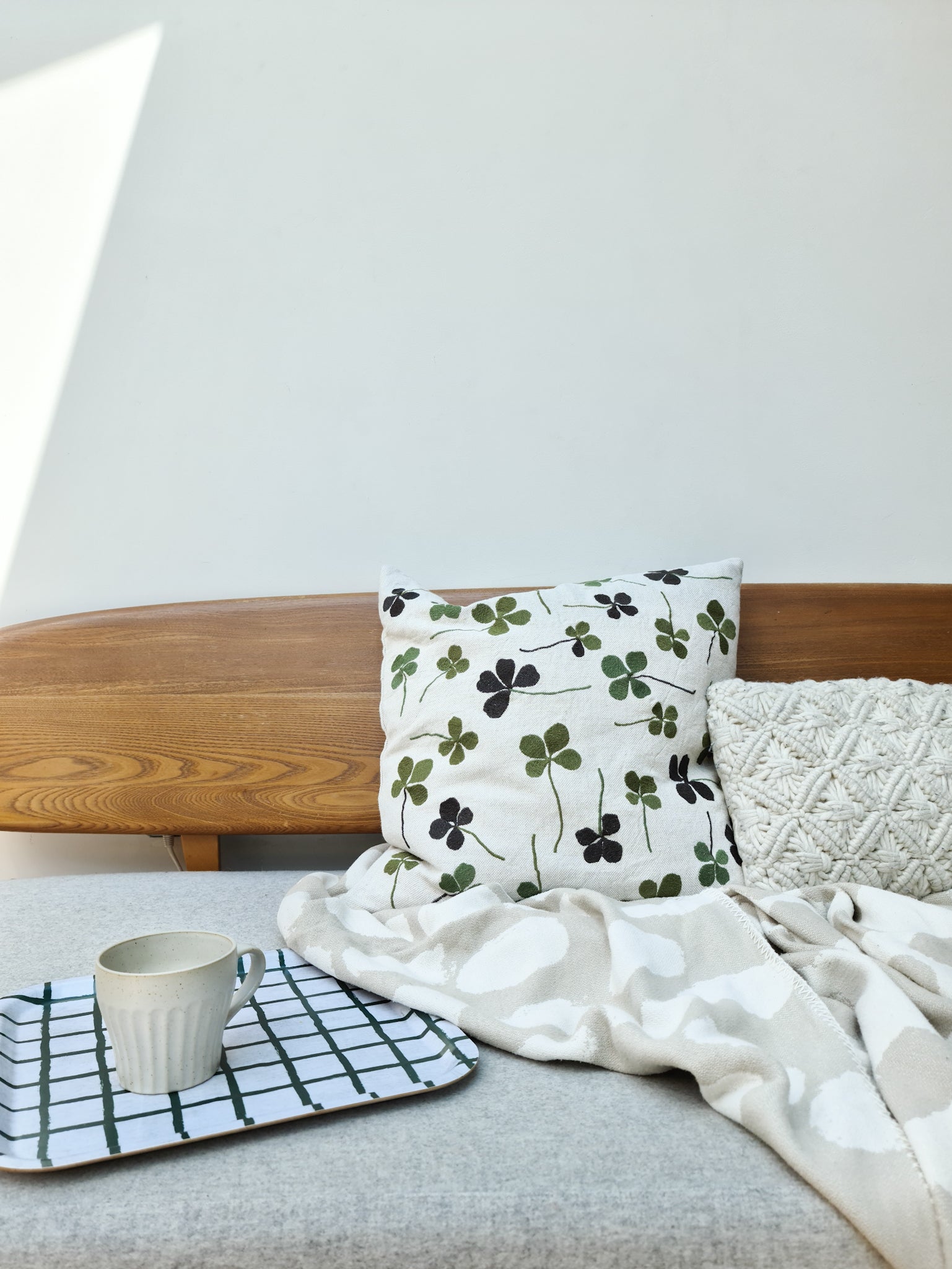 A linen coloured cushion with a green clover embroidery pattern on a sofa, next to a blanket and tray with a mug on it.