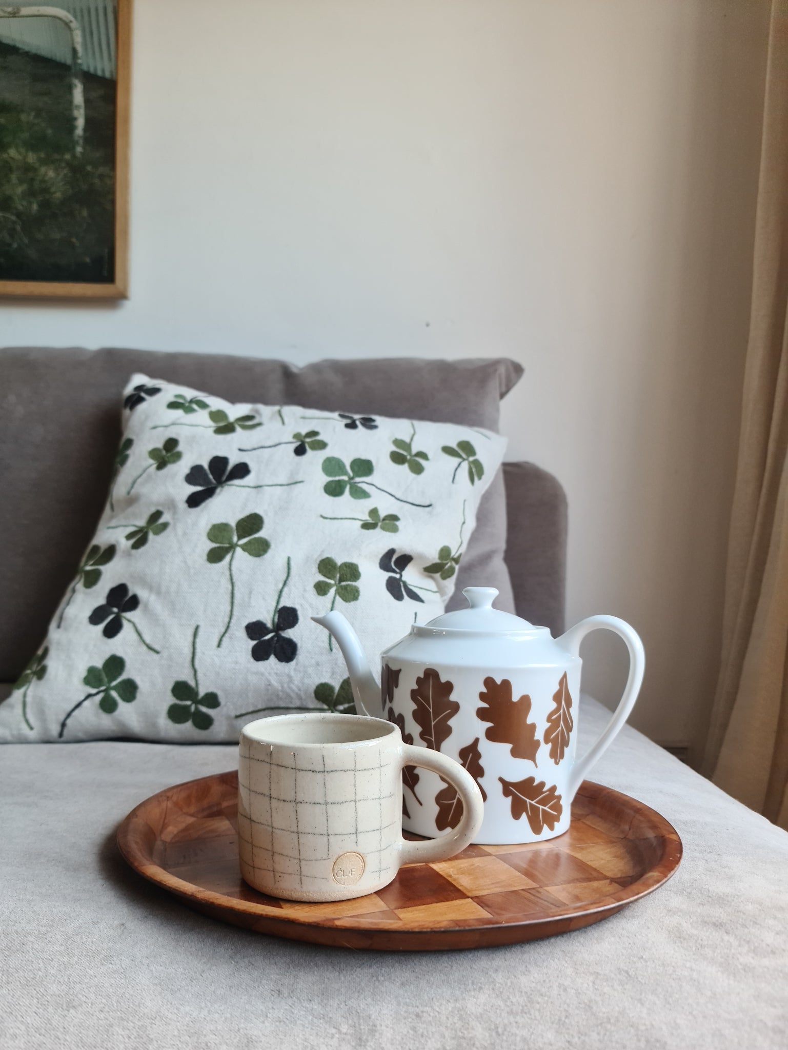 A linen coloured cushion with a green clover embroidery pattern on a sofa behind a wooden tray holding a teapot and mug on it.