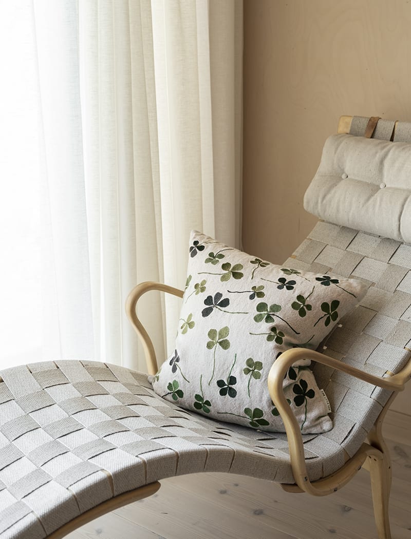 A linen coloured cushion with a green clover embroidery pattern on a reclining armchair in a neutral room.
