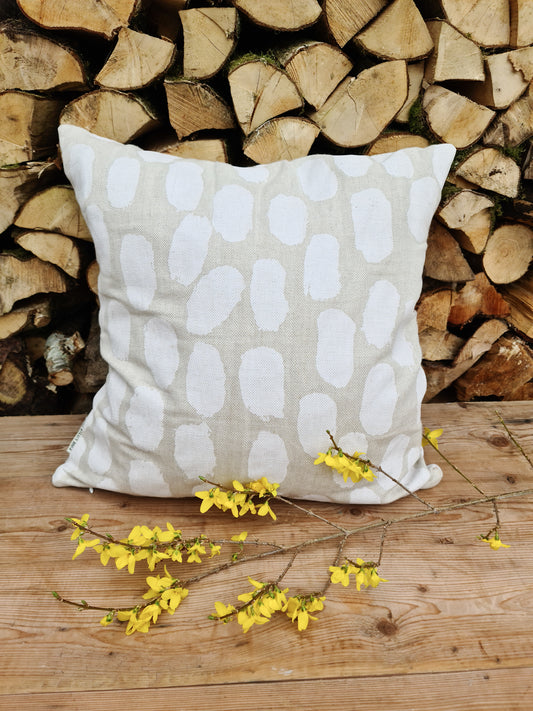 Linen cushion with a white dot screen printed pattern resting on a wooden bench against stacked wood logs and a yellow flower in-front. 