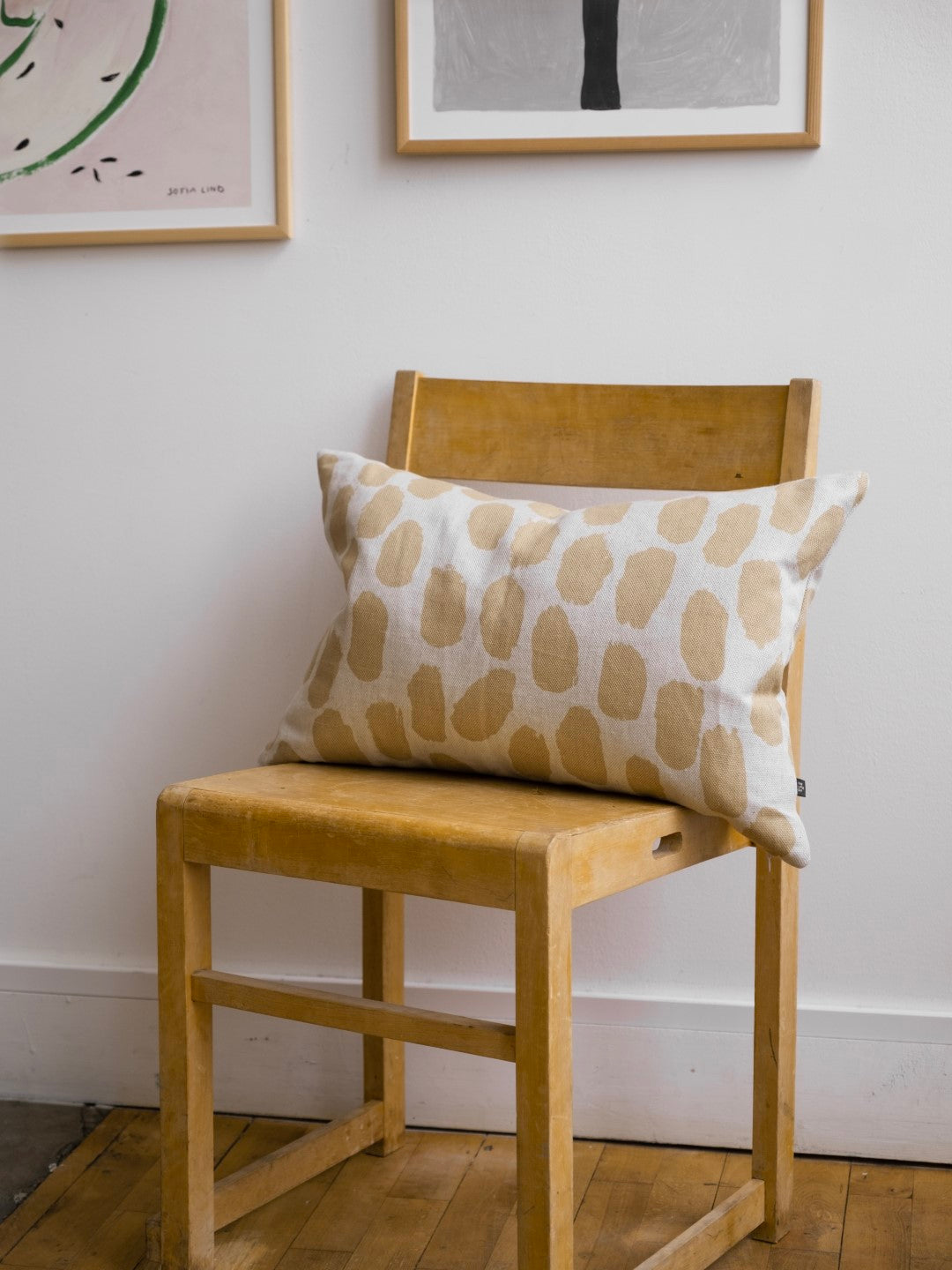 A cushion with a dotted pattern in a white and sand colourway placed on a wooden chair against a white wall.