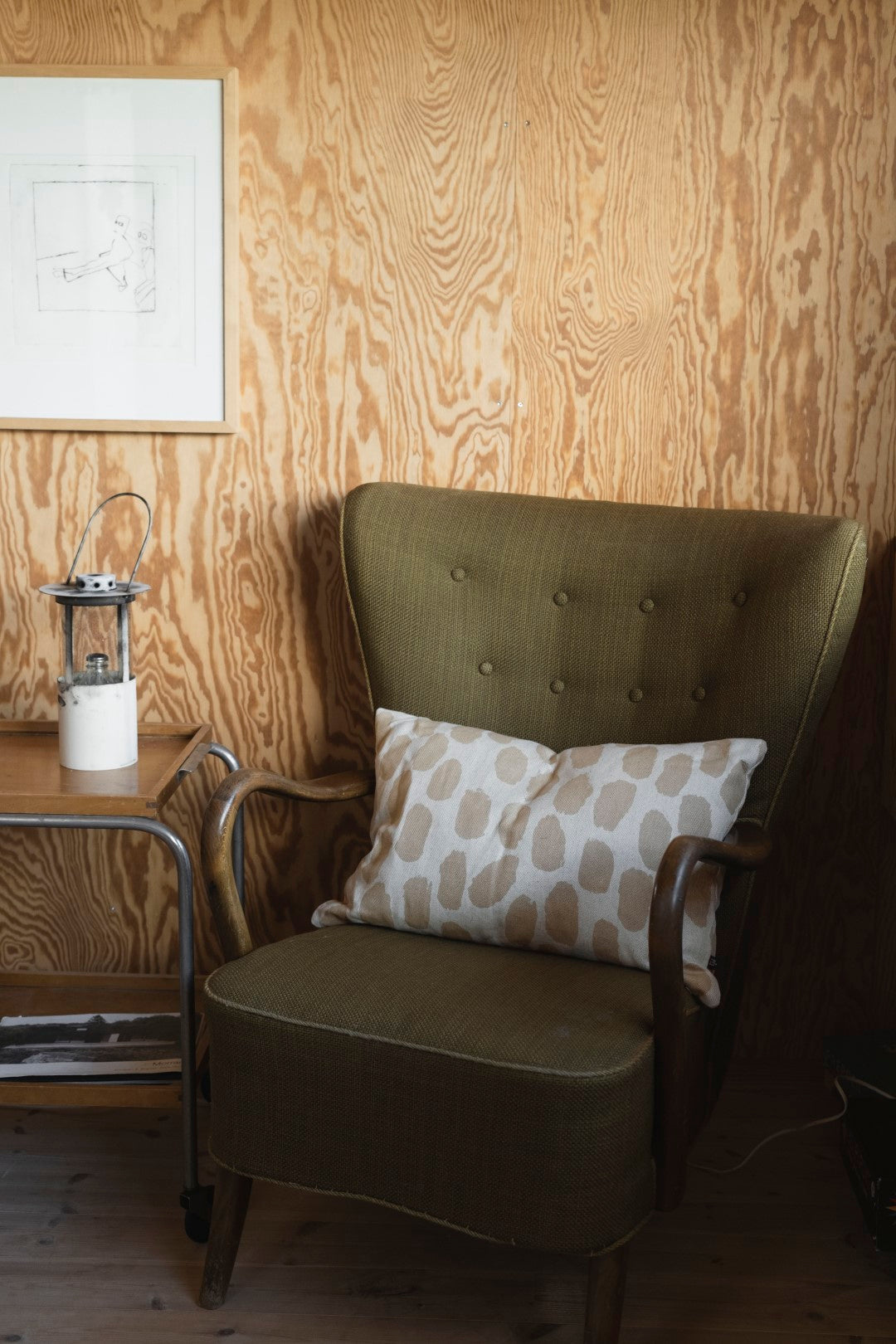 A cushion with a dotted pattern in a white and sand colourway placed on a green chair against a wooden wall.