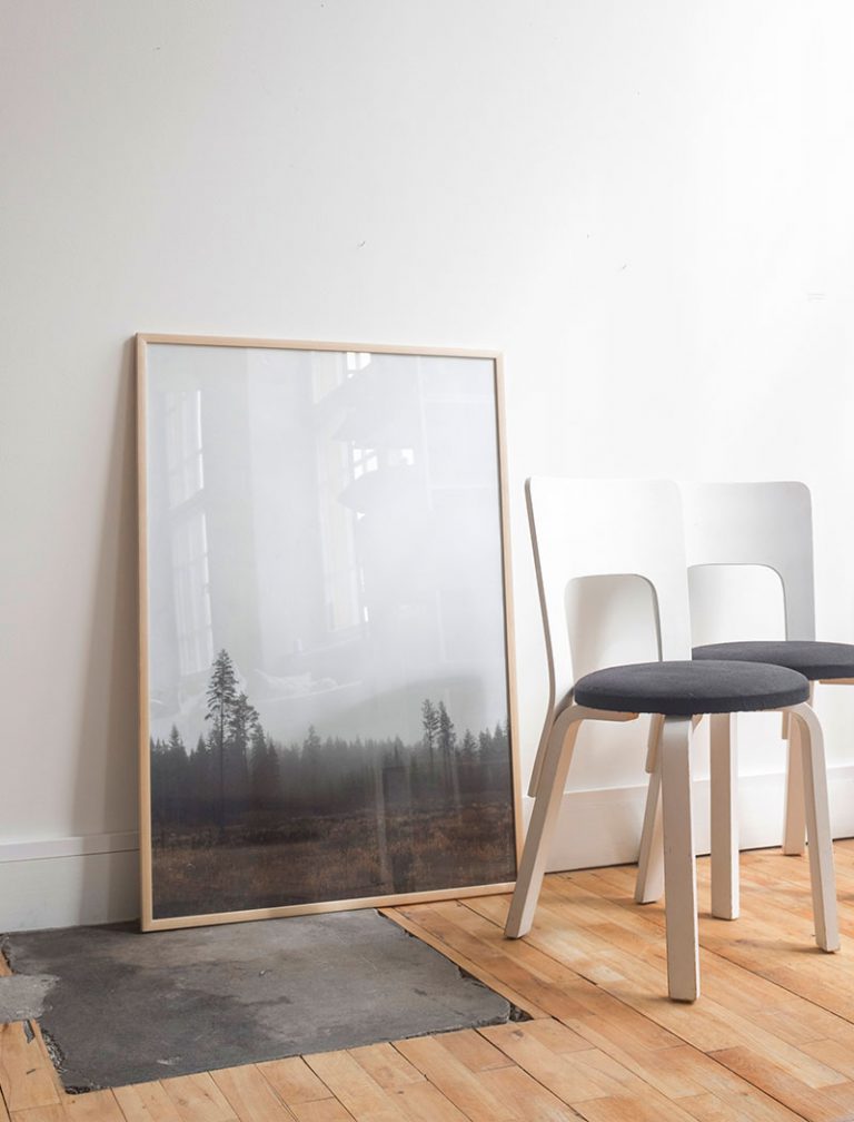 Framed poster of a Scandinavian forest propped up against a white wall, sitting on a wooden floor with two white chairs placed nearby.