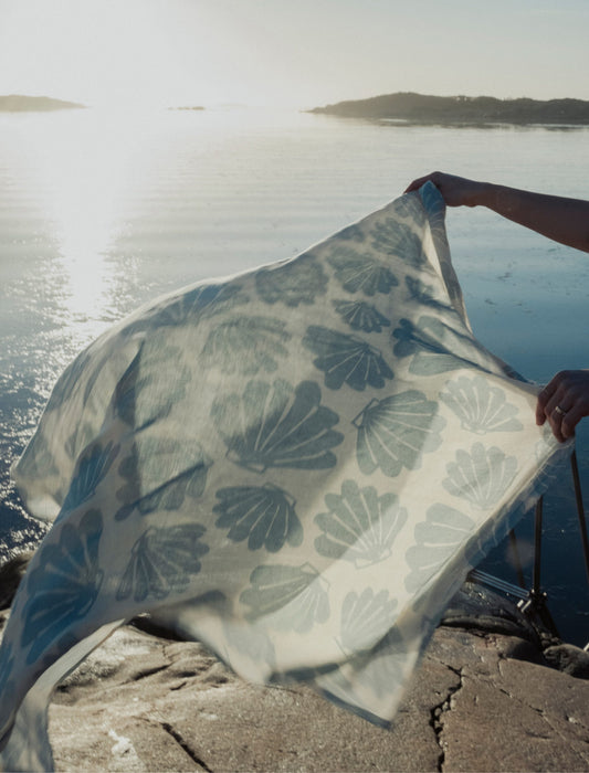 A person holding a light off-white and aqua muslin blanket with a shell pattern, by the water at sunset.