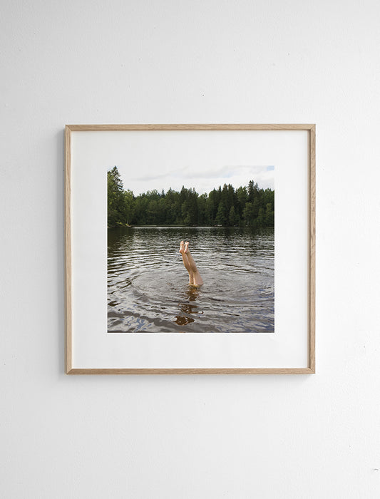 Framed photograph of a woman's legs sticking out of the water of a lake, with the water rippling around her and trees in the background.