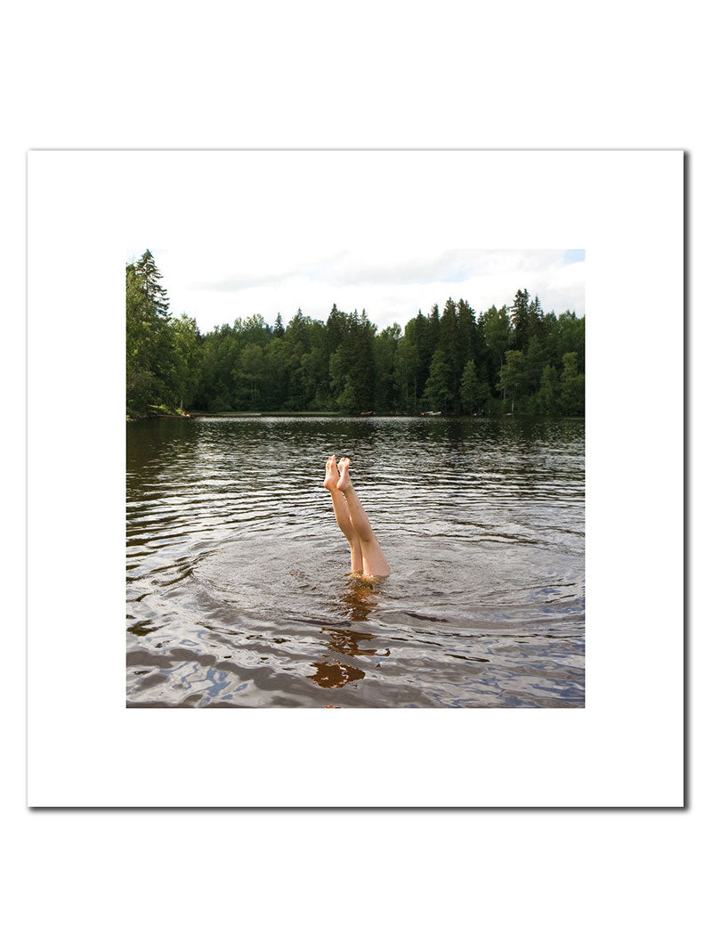 Framed photograph of a woman's legs sticking out of the water of a lake, with the water rippling around her and trees in the background.