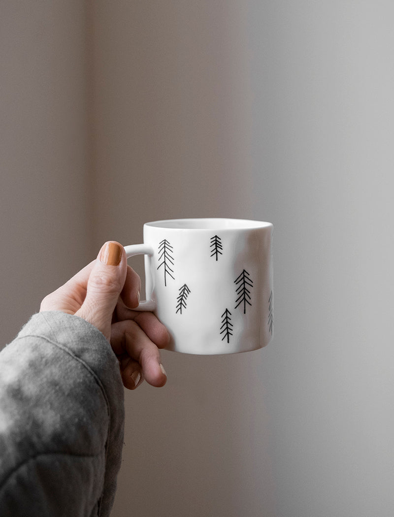 A person holding up a white porcelain cup with a pine tree motif pattern against a light background.