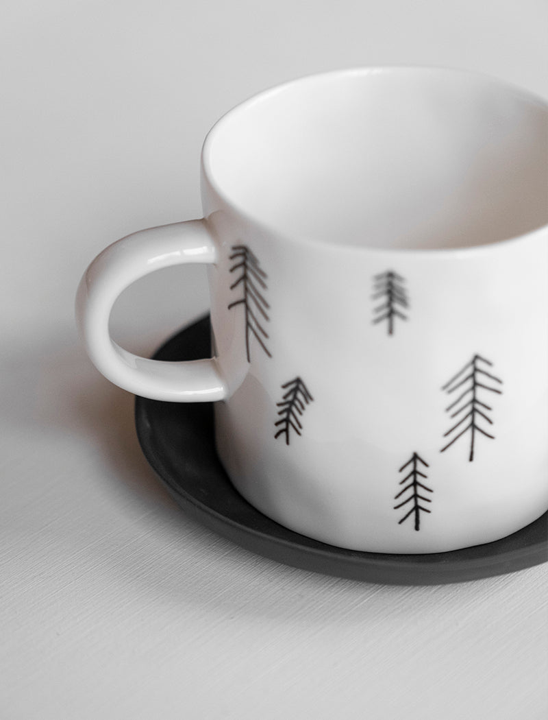 Close up of a white porcelain cup with a pine tree motif pattern against a light background.